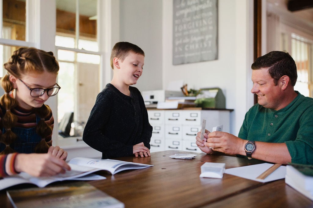 Homeschoolers practice math indoors. A girl studies from her booklet. A boy practices with flash cards.